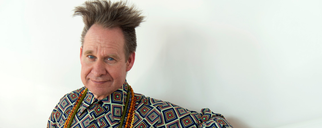 Photo of Peter Sellars in a square pattered collared shirt in front of a white background. 