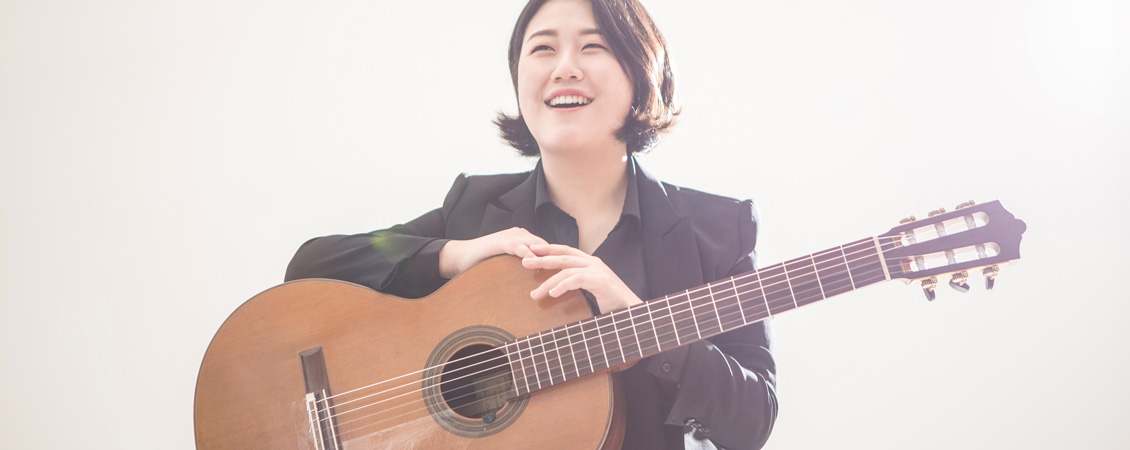 A bright photo of Bokyung in black suit holding a guitar side way looking far in the distance, smiling.