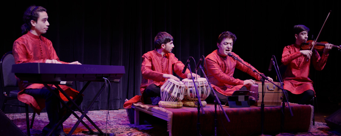 Photo of 4 guys on stage. All of them are wearing long red tops. On the left, one person is playing the keyboard. In the middle, 2 are sitting on stage with their legs criss crossed. They have microphones in front of these, one person is playing a type of drums and the other person has a wooden box in front of him. On the right, the person is playing the violin