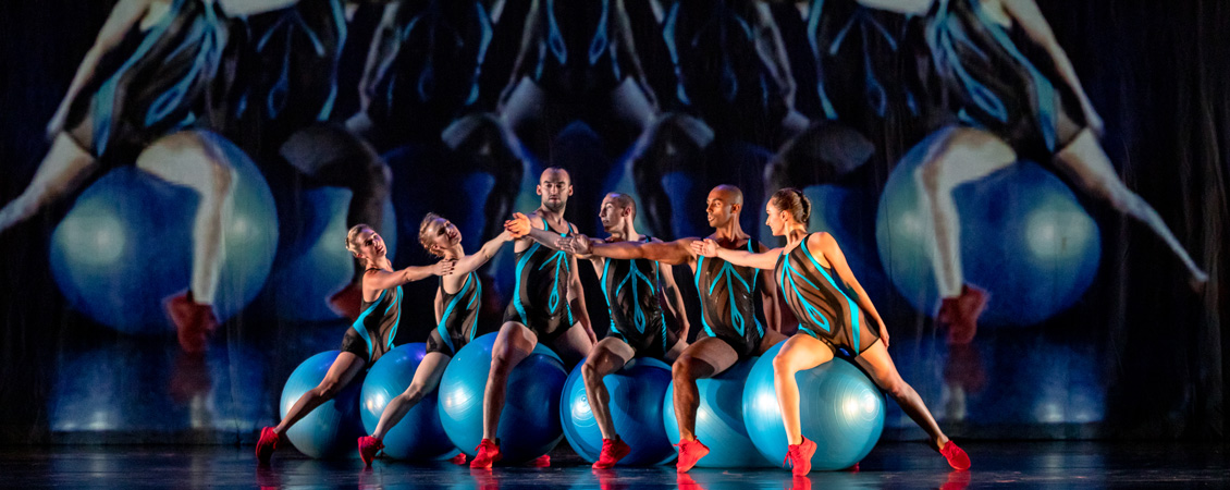An image of 6 people all wearing the same blue and black clothes with mesh material each sitting on a blue yoga ball. 