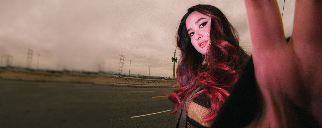 A pink sky background and a woman with long reddish hair and dramatic eyeliner