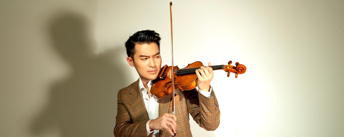 An image of a man in a tan, check patterned suit with a white collared shirt on the inside. He is playing the violine