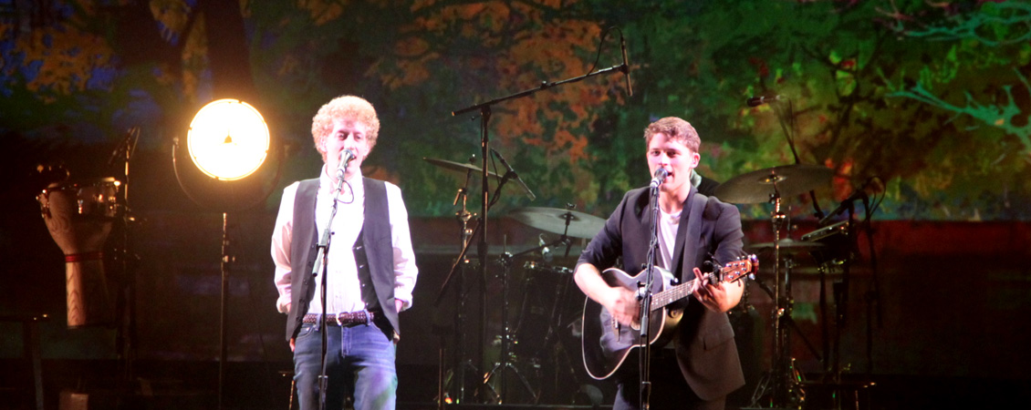 Photo of 2 guys on stage, both with microphones in front of them. Behind them there is a drum set and a light. The guy on the right is playing a guitar. 