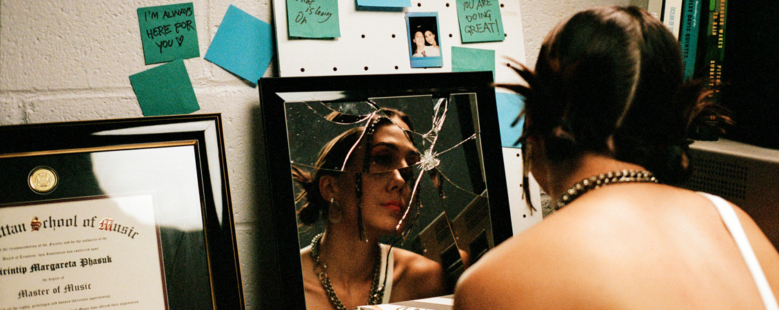 An image of Sirintip looking into a fractured mirror. The mirror is against a wall and there are sticky notes scattered along the wall. To the left of the mirror there is her diploma. 