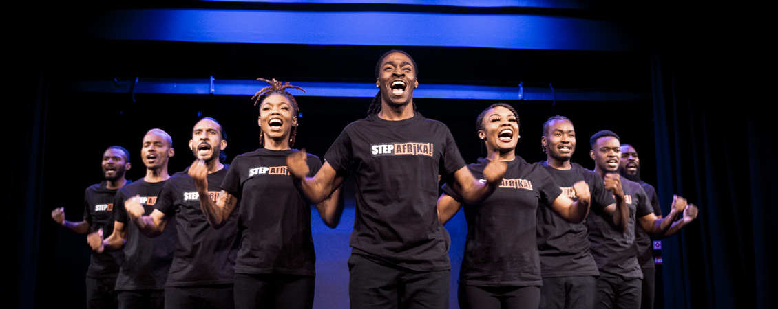 a group of people looking straight ahead in a formation of a "V", hands in fists raised to their sides and wearing Step Afrika! shirts.