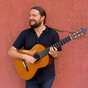 An image of a bearded man smiling and looking to the left, with a guitar in hand, against a salmon pink backdrop.