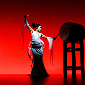 Photo of a woman standing on stage with a drum. The background/lighting of the stage is red. She is wearing a sleeveless, long white dress with pieces of cloth on the end of each of her arms. Her left hand if up in the air and right hand is hitting the drum
