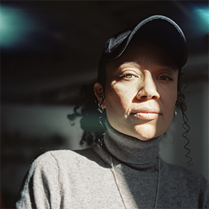 an image of GeoVanna Gonzalez in a grey turtleneck and black cap looking into the camera