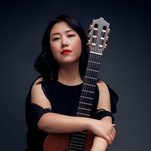 A photo of Bokyung in a black blouse holding a guitar upright