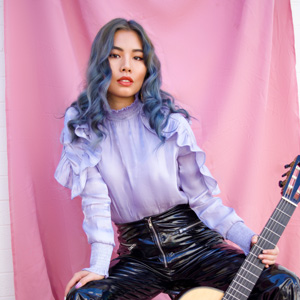 A photo of JIJI in a light purple blouse and black leather pants. There is a pink backdrop and she is holding a guitar by her legs