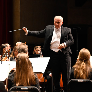 A photo of a person in white shirt and black suits conducting an orchestra. 