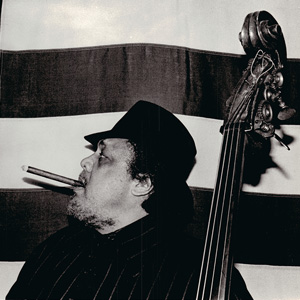 A black and white image of Charles Mingus with his bass in his hand and a cigar in his mouth.
