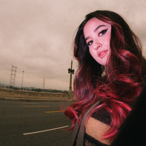 A pink sky background and a woman with long reddish hair and dramatic eyeliner