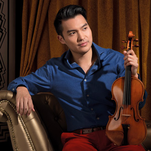 An image of a man in a blue collared shirt and red pants sitting on a bronze chair in front of brown curtains. He is looking to the right and holding a violin with his right hand 