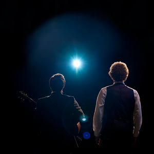 Photo of 2 people taken behind them. It seems like they are on stage, person on the left is holding a guitar and we see the stage light shining at them in the middle