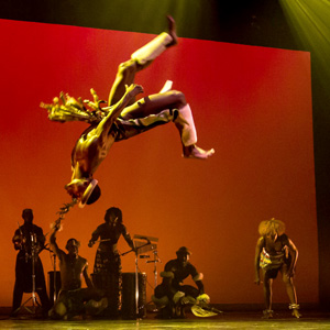 A man doing a flip in mid air while a group of people stand in anticipation in the background. The stage is lit a sunset orange.