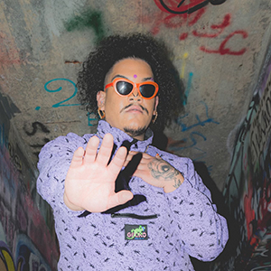 an image of XayRoZay with neon-orange lined sunglasses and a high-collared purple shirt, with one hand on his chest and the other extended out