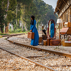 An image of a woman in a blue dress looking down. She stands next to train tracks with trunks and miscellaneous furnitures by her side.