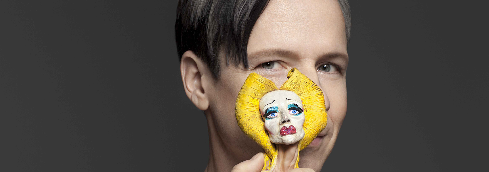 John Cameron Mitchell is pictured with a figurine in front of his face. The figurine is in heavy makeup with golden hair.