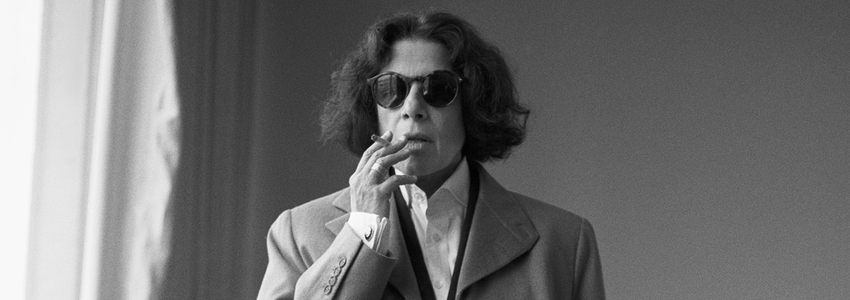 Fran Lebowitz is pictured with shoulder-length hair, sunglasses, and a suit jacket smoking a cigarette in a black and white photo.