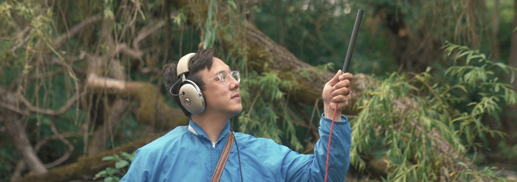 No-No Boy is pictured standing in the woods wearing headphones and holding a microphone.