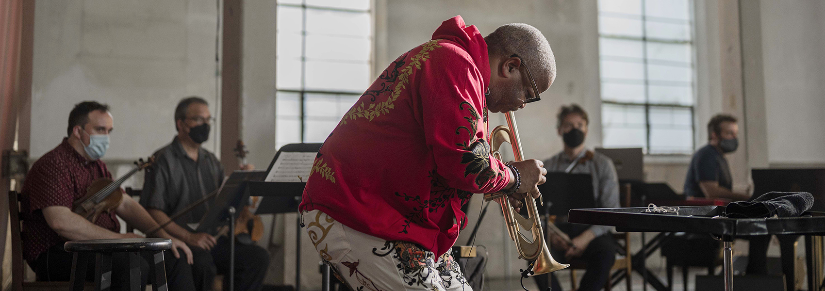 Terrence Blanchard wears a vibrant red and gold jacket plays the trumpet passionately, with a group of masked musicians sitting behind him in a rehearsal space.