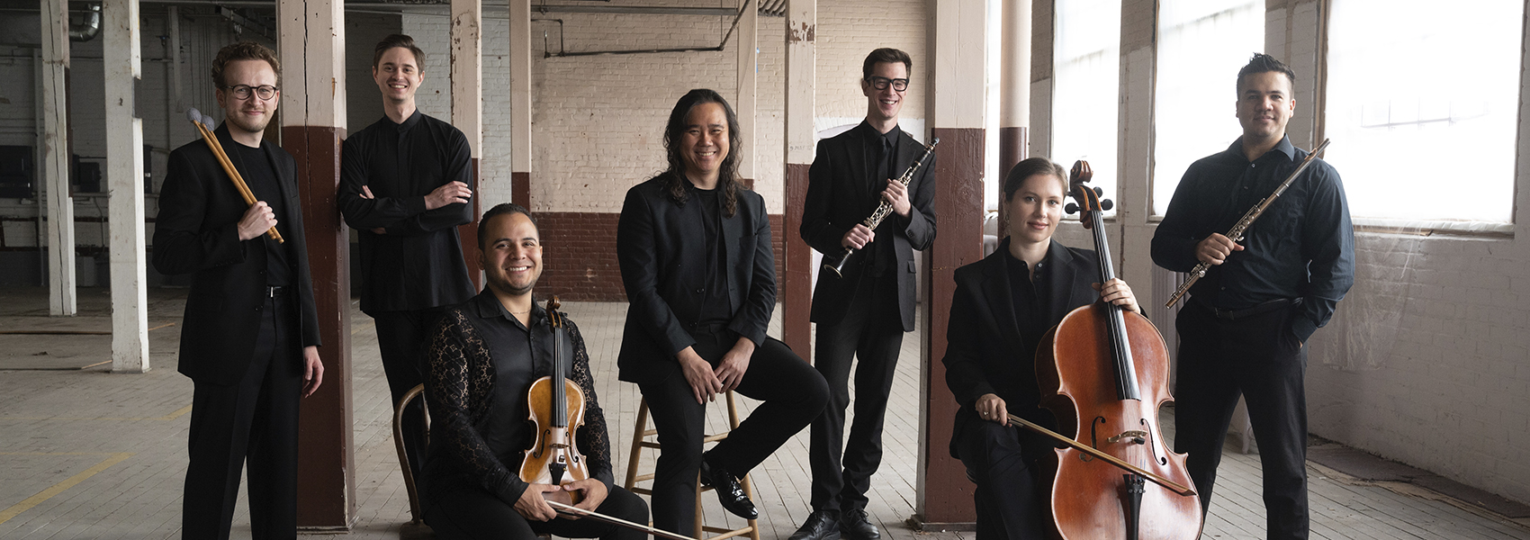 The musicians, dressed in black and holding their instruments, pose together in a sunlit room with large windows.