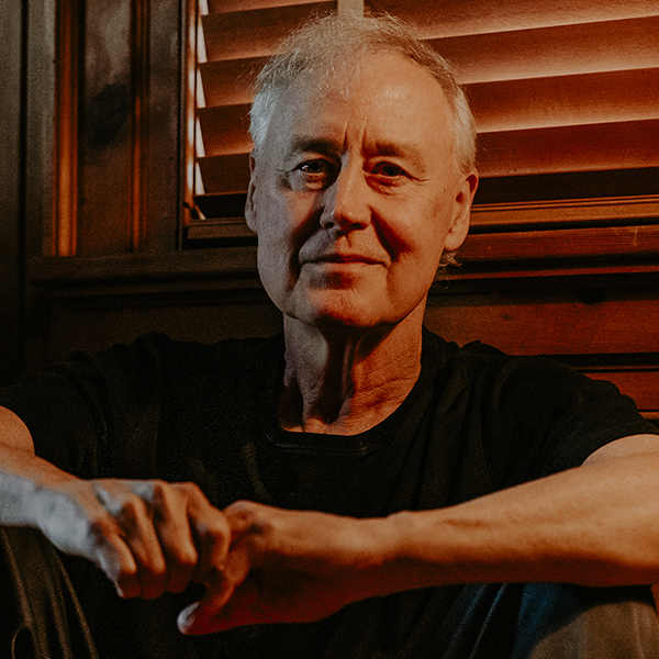 Bruce Hornsby sits on the floor with his arms on his knees next to a dollhouse.