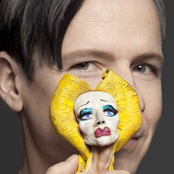 John Cameron Mitchell is pictured with a figurine in front of his face. The figurine is in heavy makeup with golden hair.