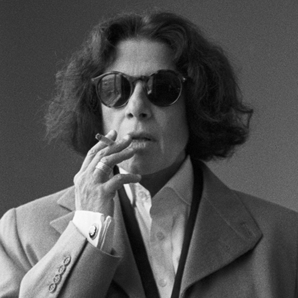 Fran Lebowitz is pictured with shoulder-length hair, sunglasses, and a suit jacket smoking a cigarette in a black and white photo.