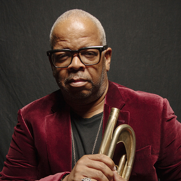 Terrence Blanchard wears a vibrant red and gold jacket plays the trumpet passionately, with a group of masked musicians sitting behind him in a rehearsal space.