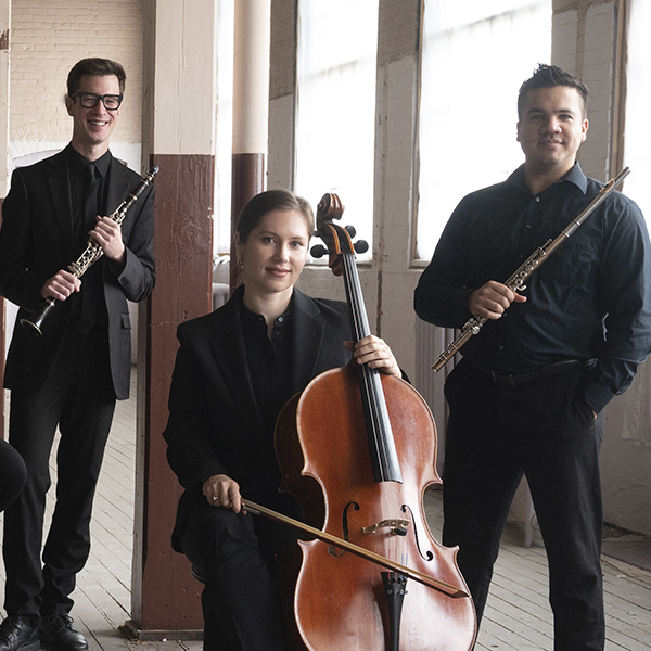 The musicians, dressed in black and holding their instruments, pose together in a sunlit room with large windows.