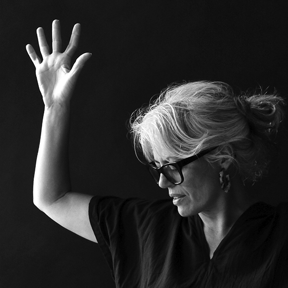 Yanira Castro stands with her hands raised in a dynamic pose, set against a black background, captured in black and white.