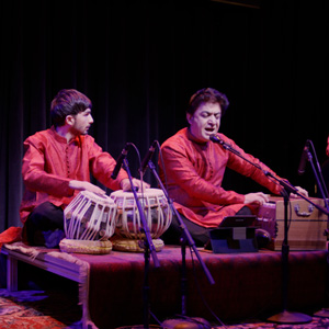 Photo of 2 guys sitting on stage with their legs criss crossed. They are wearing long red tops. They have microphones in front of these, one person is playing a type of drums and the other person has a wooden box in front of him.