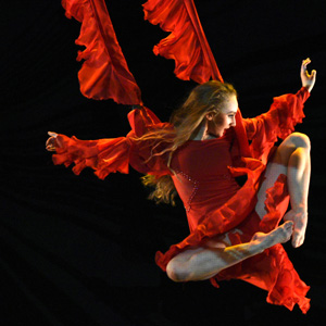 An image of a woman in red dress with flowy long sleeves, jumping in the air with her legs tucked below her.