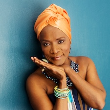 Angelique Kidjo looking at the camera in an orange turban against a teal background